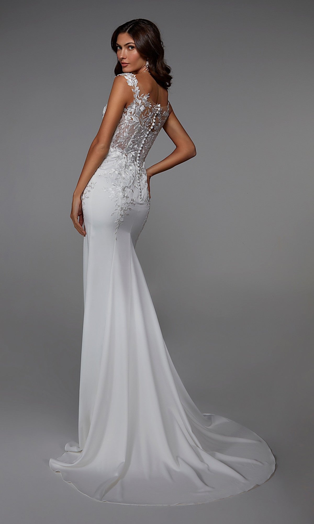 A woman models the Long White Dress: Alyce 7026, featuring a fitted lace design with a sheer floral back, as she stands against a plain gray background looking over her shoulder.